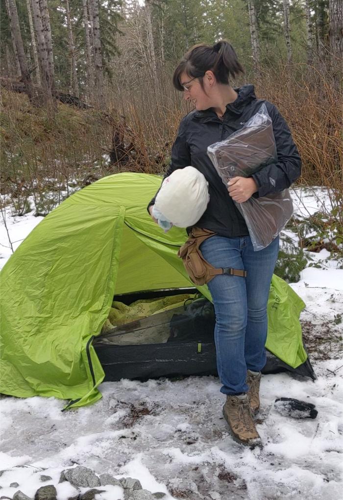 Raffin stands, camping equipment in her arms, outside of a camping tent in a snowy forest.
